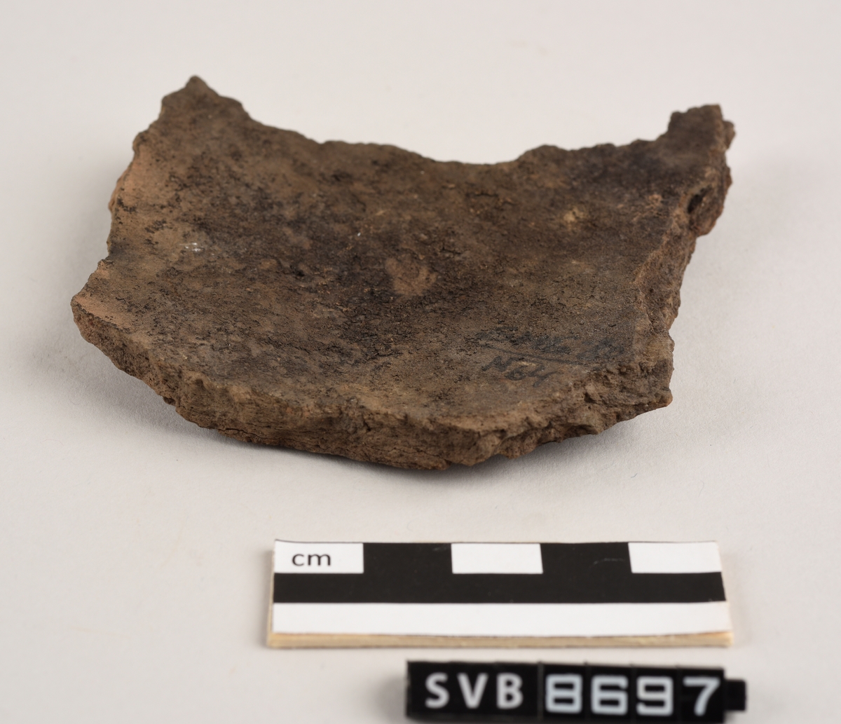 Lett buet keramikkfragment med bruddkanter på alle sidene. Fragmentet er brunt i farge. Bruddkantene er brune i farge.