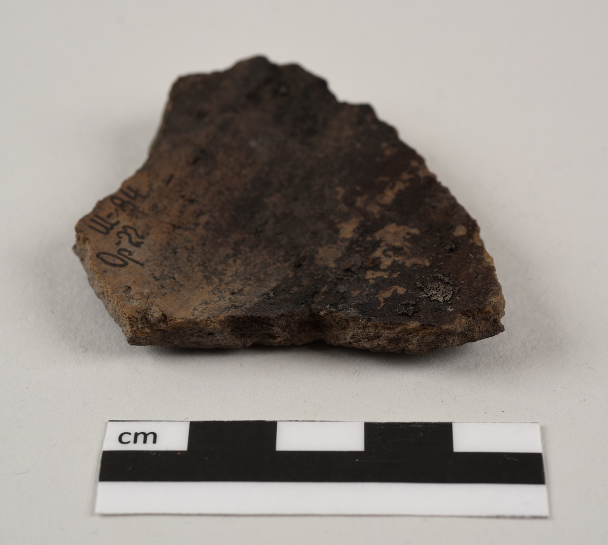 Lett buet keramikkfragment med bruddkanter på alle sidene. Fragmentet er mørkebrunt til svart i farge. Bruddkantene er grå i farge.
