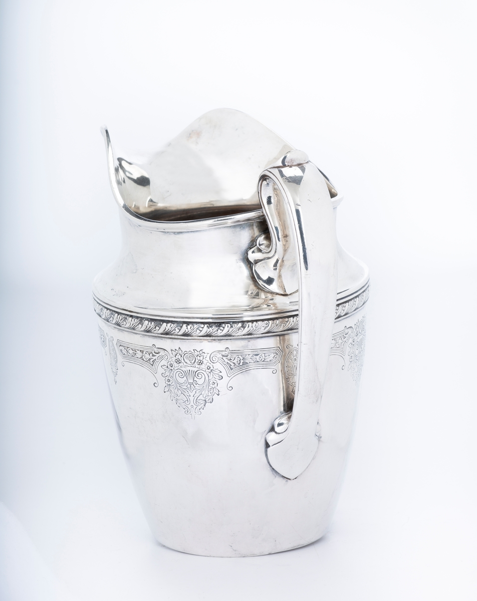 Mugge, blankt metall, bred tut, slank hank. Smal bord rundt muggen med høyt relieff, derunder siselerte blomsterrosetter, bånd og medaljonger. Stempel:? Under bunden er innrisset JVB. 