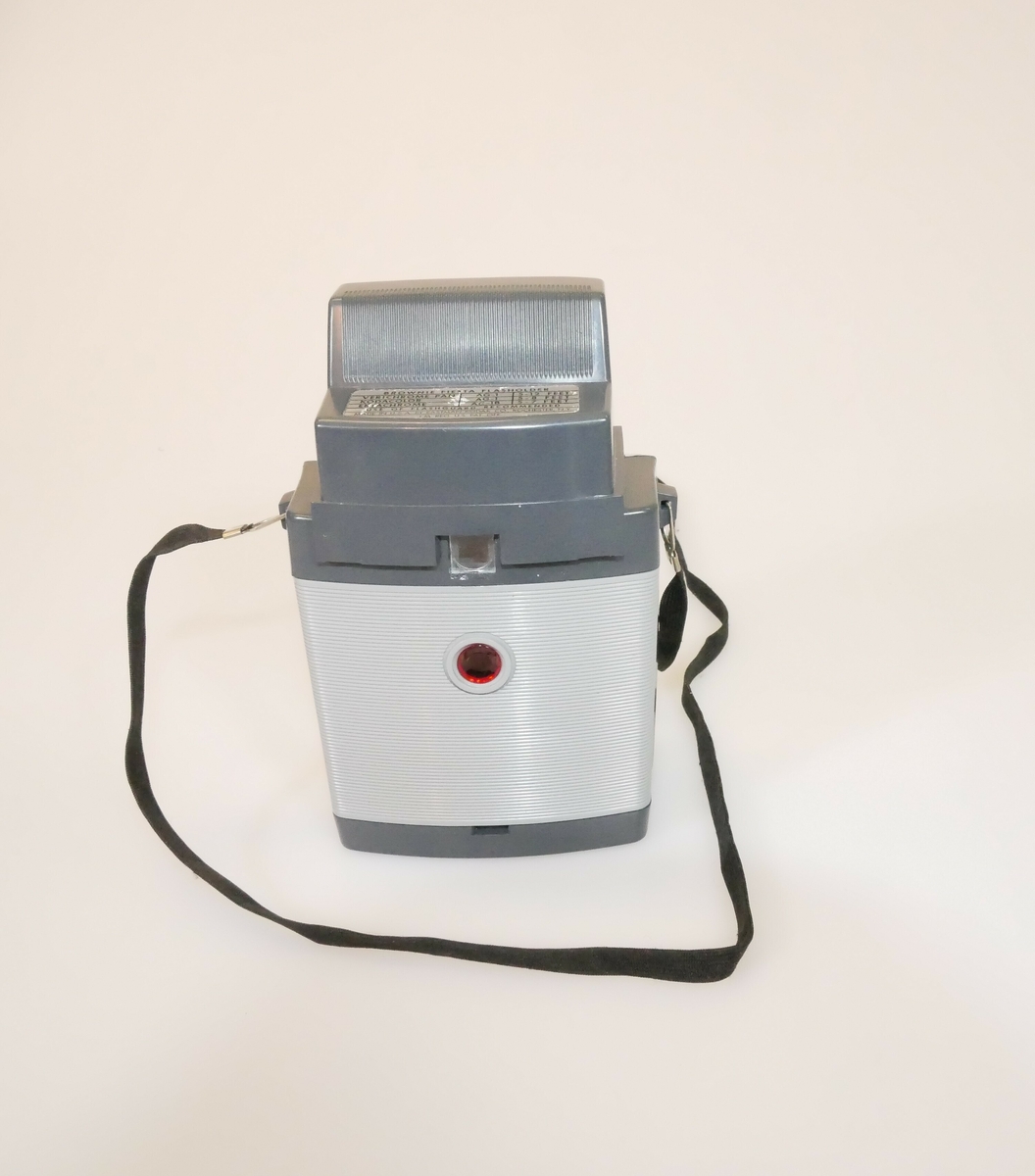 Rektangulært fotoapparat. Lysegrå forside dekket av klar hard plast, midt på er det en linse. På toppen er det boks av grå hard plast til blits. På baksiden er det grå og lysegrå hard plast. Sort bånd festet på begge sider av kameraet med metallfeste. 