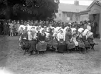 Svenskbybor på Ryhovs kasernområde i Jönköping 1929. Kvinnor och barn på bänkar vid lördagens gudstjänst med biskop Edgar Reuterskiöld. Pojken längst till vänster har fått bandage om några fingrar. Kvinnan nr 5 från höger som tar handen i sin dotters hår heter Melitta Knutas född Felsing dottern heter Adelina.