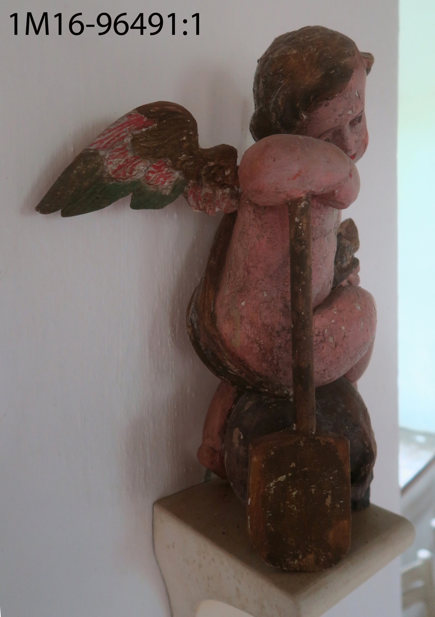 Skulpterad ängel av trä. Sitter på dödskalle med huvudet något högerböjt. Håller en snäcka med tre frukter (?) i höger hand. Draperat tygstycke runt midjan, över höger axel och rygg, avslutas framtill. Saknar vingar, utsträckningar i skuldrorna visar var de suttit. Vänster arm och höger fot saknas. Har hållit ett nu avbrutet föremål i munnen. Träplugg i botten avbruten. Sekundär målning i mörk hudfärg med röda kinder. Hår, dödskalle och övriga detaljer i brunt. Färgen fläckvis borta. Har troligen tillhört epitafium eller altaruppsats.