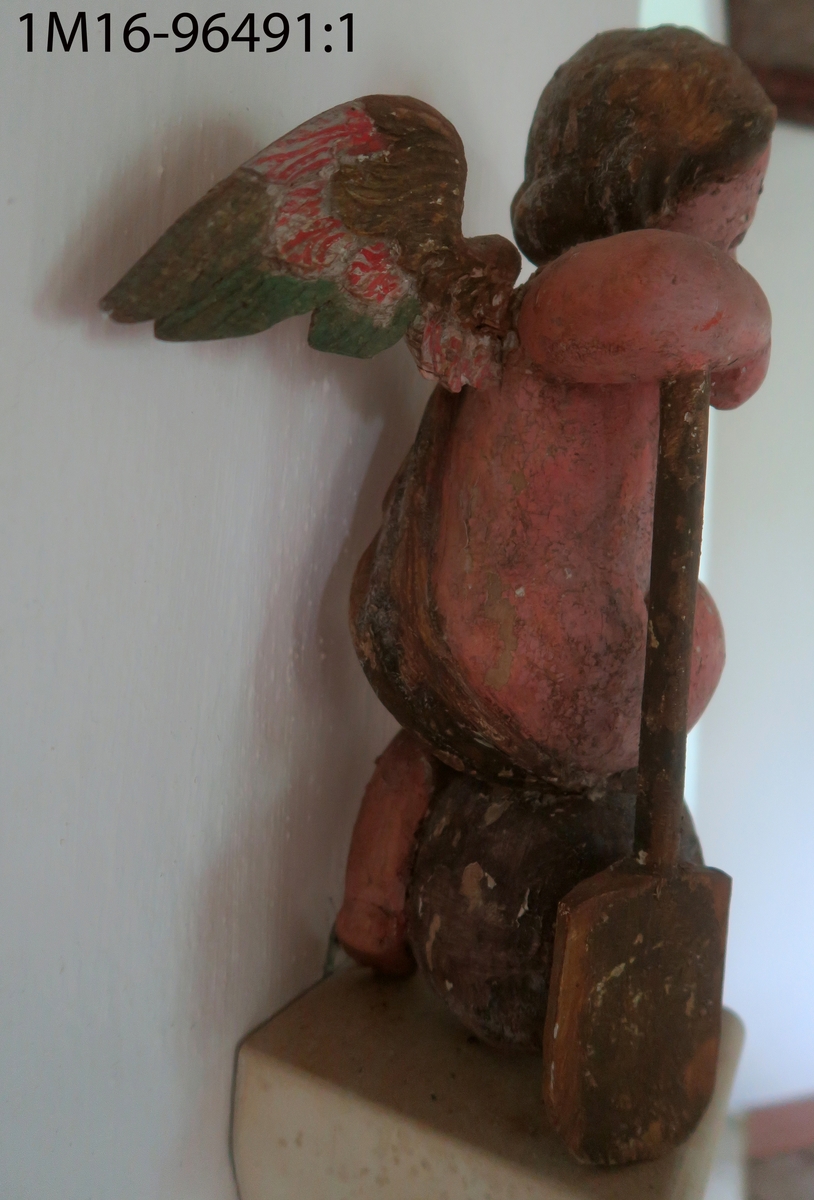 Skulpterad ängel av trä. Sitter på dödskalle med huvudet något högerböjt. Håller en snäcka med tre frukter (?) i höger hand. Draperat tygstycke runt midjan, över höger axel och rygg, avslutas framtill. Saknar vingar, utsträckningar i skuldrorna visar var de suttit. Vänster arm och höger fot saknas. Har hållit ett nu avbrutet föremål i munnen. Träplugg i botten avbruten. Sekundär målning i mörk hudfärg med röda kinder. Hår, dödskalle och övriga detaljer i brunt. Färgen fläckvis borta. Har troligen tillhört epitafium eller altaruppsats.