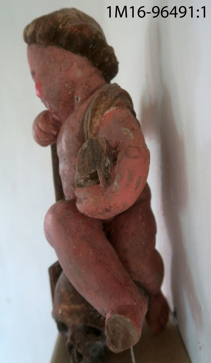 Skulpterad ängel av trä. Sitter på dödskalle med huvudet något högerböjt. Håller en snäcka med tre frukter (?) i höger hand. Draperat tygstycke runt midjan, över höger axel och rygg, avslutas framtill. Saknar vingar, utsträckningar i skuldrorna visar var de suttit. Vänster arm och höger fot saknas. Har hållit ett nu avbrutet föremål i munnen. Träplugg i botten avbruten. Sekundär målning i mörk hudfärg med röda kinder. Hår, dödskalle och övriga detaljer i brunt. Färgen fläckvis borta. Har troligen tillhört epitafium eller altaruppsats.