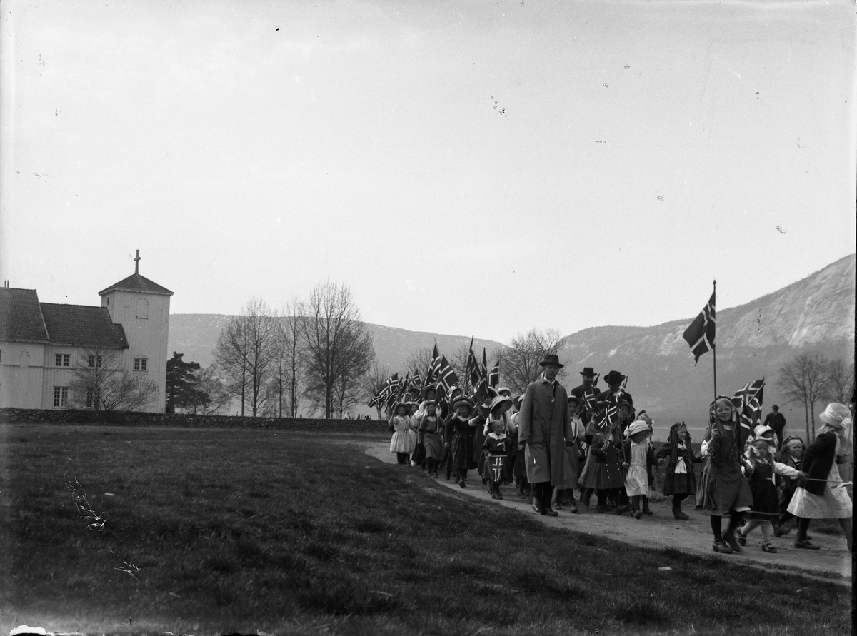 Fotosamling etter Bendik Ketilson Taraldlien (1863-1951) Fyresdal. Gårdbruker, fotograf og skogbruksmann. Fotosamlingen etter fotograf Taraldlien dokumenterer områdene Fyresdal og omegn. 
Motiv med 17. maitog forbi Moland kirke i Fyresdal.