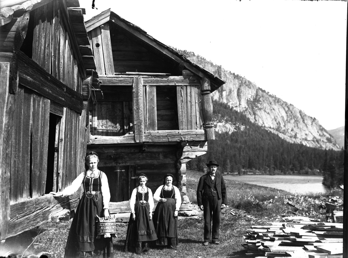 Fotosamling etter Bendik Ketilson Taraldlien (1863-1951) Fyresdal. Gårdbruker, fotograf og skogbruksmann. Fotosamlingen etter fotograf Taraldlien dokumenterer områdene Fyresdal og omegn. 
Portrett av K. Snarteland m/familie ved stabbura på Snarteland. På det ene stabburet finnes runer fra 1200 eller 1300 tallet på dørhengslet.