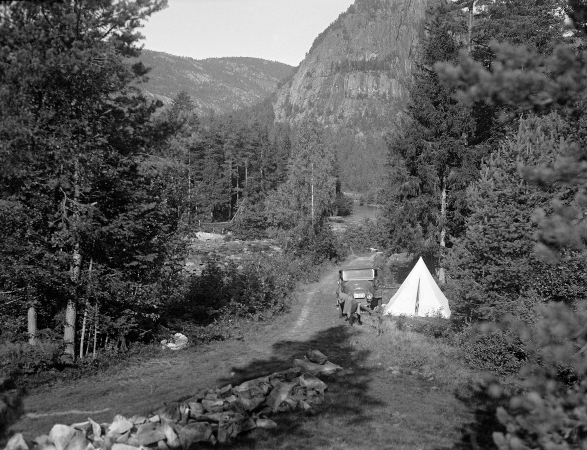 Fra familien Midttuns ferietur sommeren 1933.  Fotografiet er tatt fra en bakke der der gikk en grasbevokst sideveg.  Nederst i bakken hadde familien parkert bilen (en Opel fra 1920) og slått opp det kvite pyramideteltet de skulle overnatte i på en liten grasflekk.  I bakgrunnen skimter vi et elveløp omgitt av furudominert skog og til dels også bratte bergskrenter med vegetasjon bare i revnene.  Fotografiet er tatt av bileieren, fylkesskogmester Jørun Midttun, i en av hans ferier.  Midttun arbeidet i Telemark, men hadde også familie i Hardanger.  Vi vet ikke nøyaktig hvor dette fotografiet er tatt.  Teltferie var populært i mellomkrigstida, og de forholdsvis få familiene som hadde egen bil reiste gjerne, både for å besøke familie og for å se andre deler av landet. Friluftsliv.
