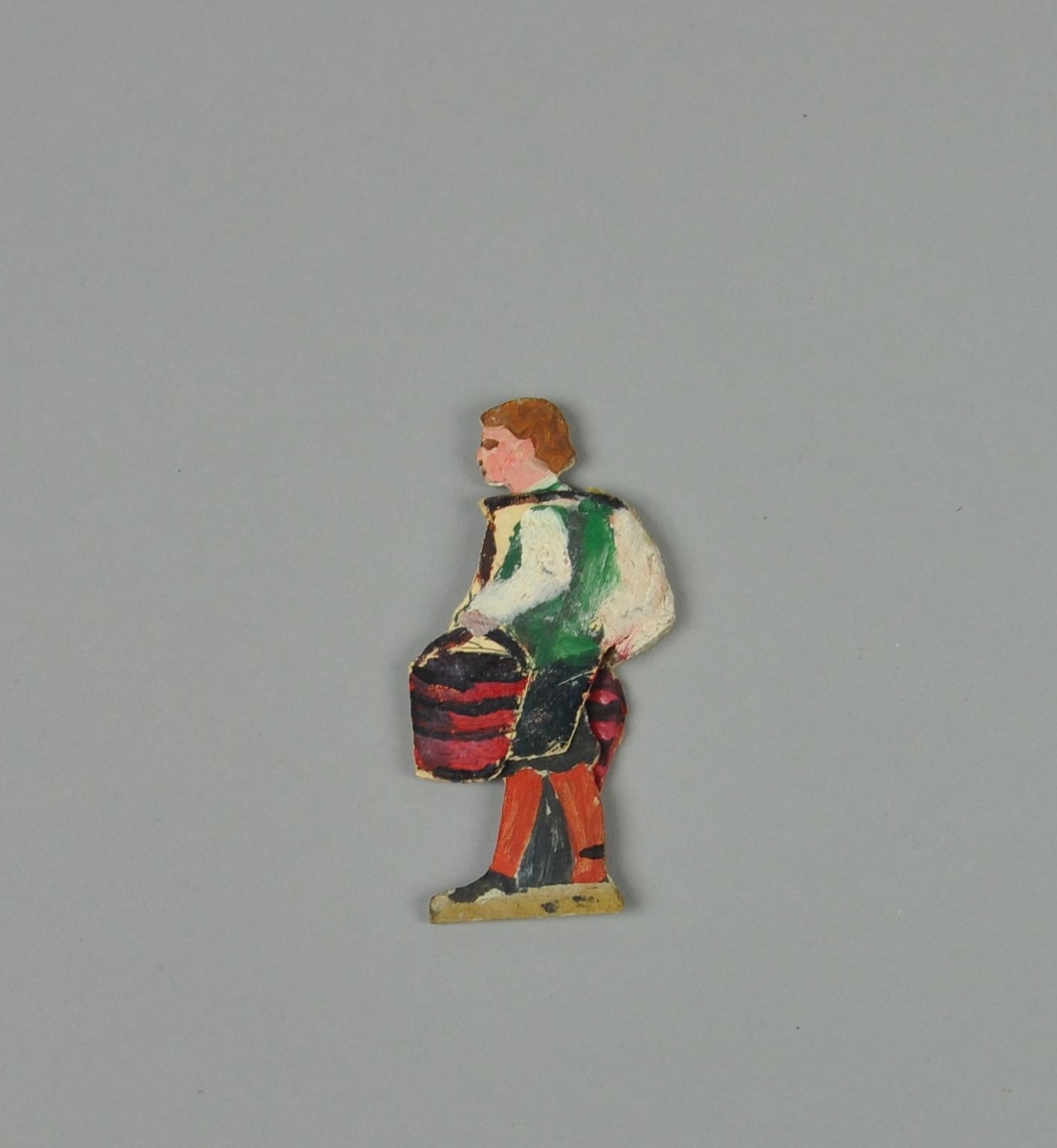 Figur av papp. Figuren forestiller gutt i sideprofil, med hendene i lommen. En annen del kan skyves over figuren, slik at gutten holder to bøtter. Figuren er malt på begge sider.