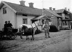 TVÅ MÄN SITTER I EN HÄSTVAGN UTANFÖR ETT BOSTADSHUS I 
TÖREBODA. DEN VITA DRAGHÄSTEN HAR ETT FÖL VID SIDAN.