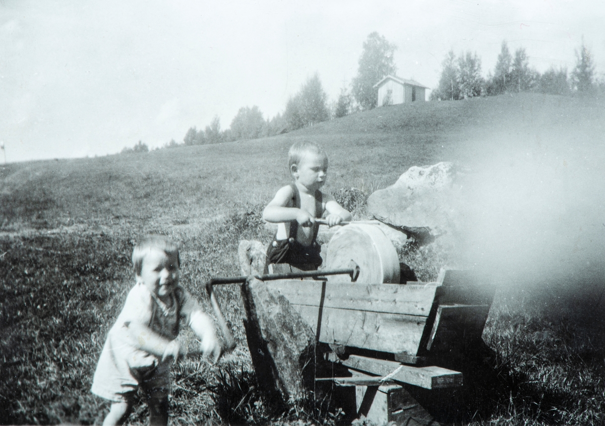 2 barn med en slipstein. Et barn sveiver på steinen, det andre sliper redskapen. Oustad?