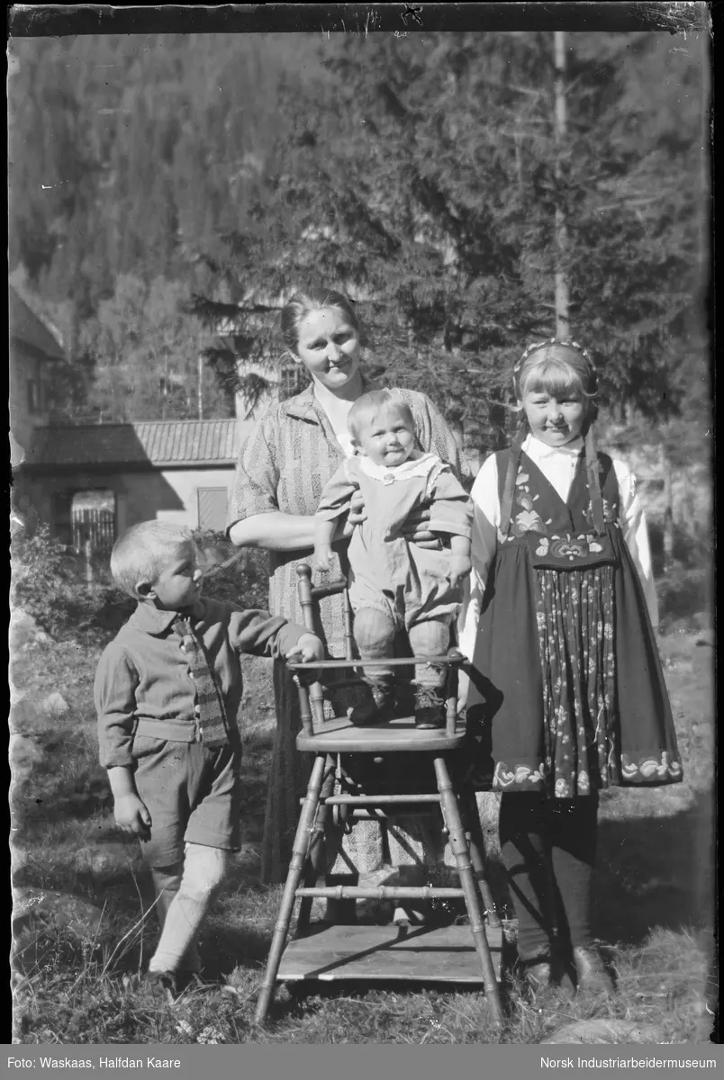 Kvinne med tre barn stående foran bolighus. Jenta i bunad (variant av Hulda Garborg bunad) og spedbarn stående i barnestol.