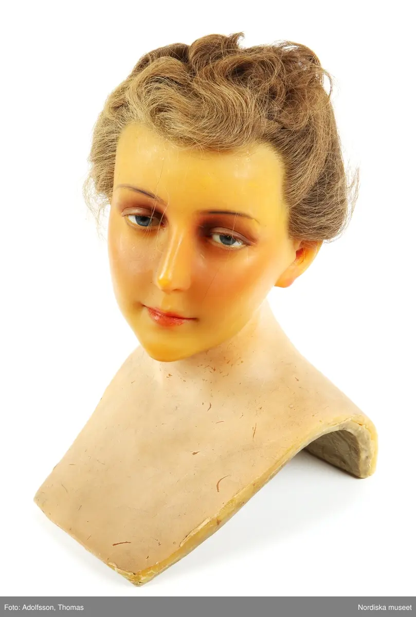 Skyltdockshuvud, kvinna, av vax med riktigt hår, ögonfransar och ögon av glas. Med hals och övre del av torso. På baksidan av torso text "Paul Baschwitz / Berlin C". Håret friserat i 1940-tals frisyr (hårnålar ingår ej i förvärvet).
Anm. spricka vid vänster öga och vax gulnat.
/Helen Persson 2018-02-13