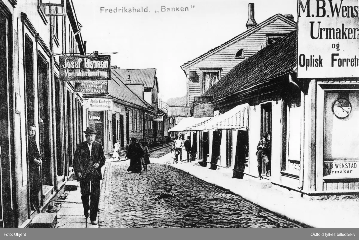Gateparti fra Halden, Banken, Nedre Bankegate ca. 1900-1910. Postkort.
Reklameskilt for urmaker M. B. Wenstad, Urmaker og optisk forretning. Eksteriør av butikken til høyre. Magnus Bernts. Wenstad, f. 1871 i Våler i Solør, urmaker. 
Til venstre forretningen til Josef Hansen, manufakturforretning. Innehaver Josef Nikolay Hansen f. 1875 i Halden. Johannesen Pølsemageri, Slagteri & Røgeri. Justine Gundersen mnaufakturforretning. Olsen.
