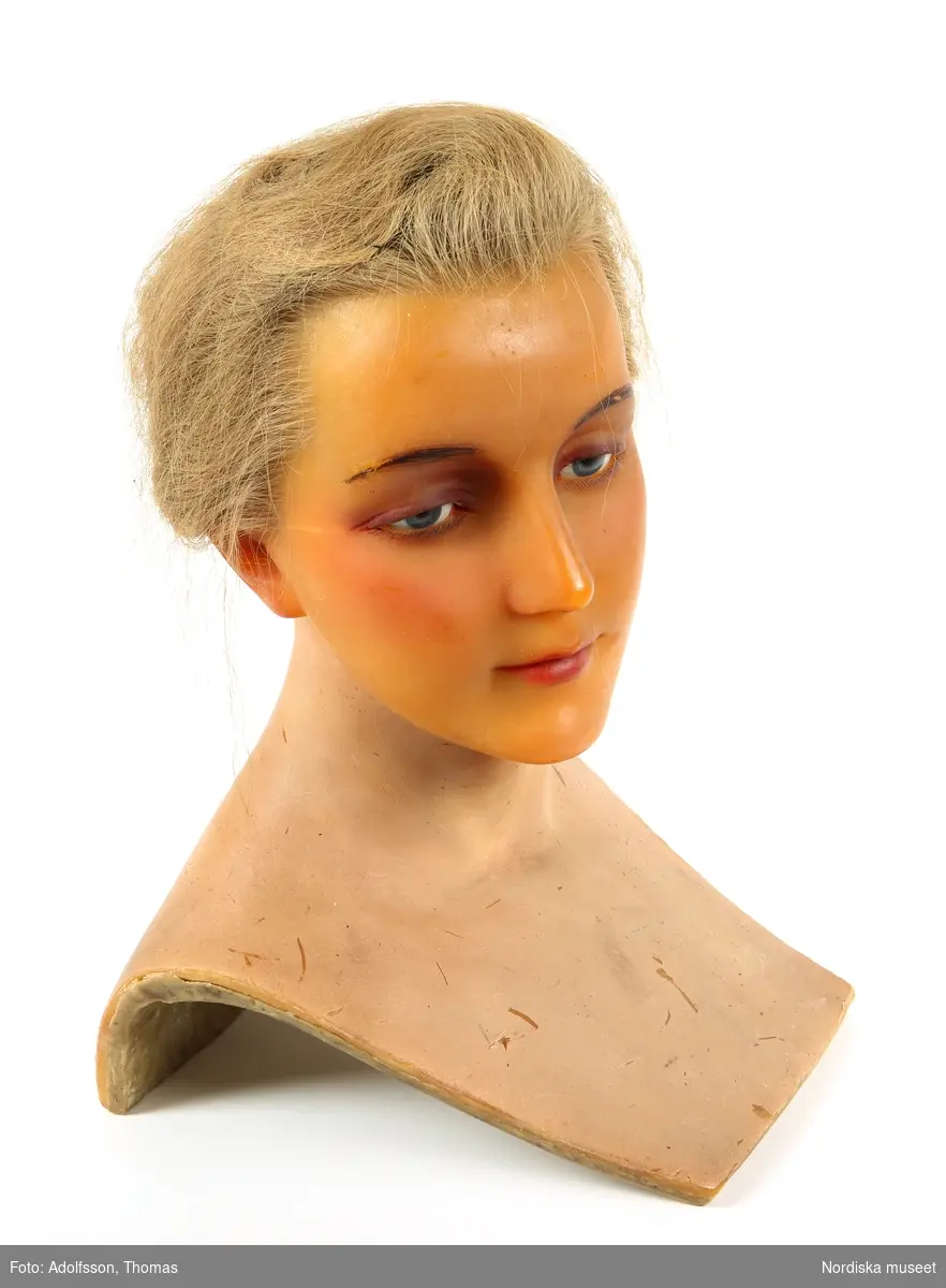 Skyltdockshuvud, kvinna, av vax med riktigt hår, ögonfransar och ögon av glas. Med hals och övre del av torso. På baksidan av torso text "Paul Baschwitz / Berlin C". Blont hår friserat i knut (hårnålar ingår ej i förvärvet).
Anm. spricka vid höger och vänster öga och vax gulnat.
/Helen Persson 2018-02-13