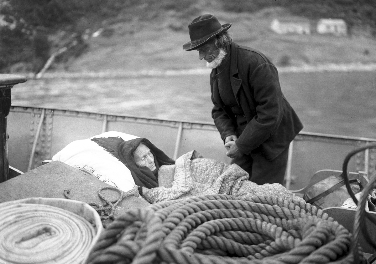 Syketransport av et eldre par i båt på vei til Ålesund. "Den syke paa Dekket". Ingeborg og Ola Hustad, Hjørundfjord.