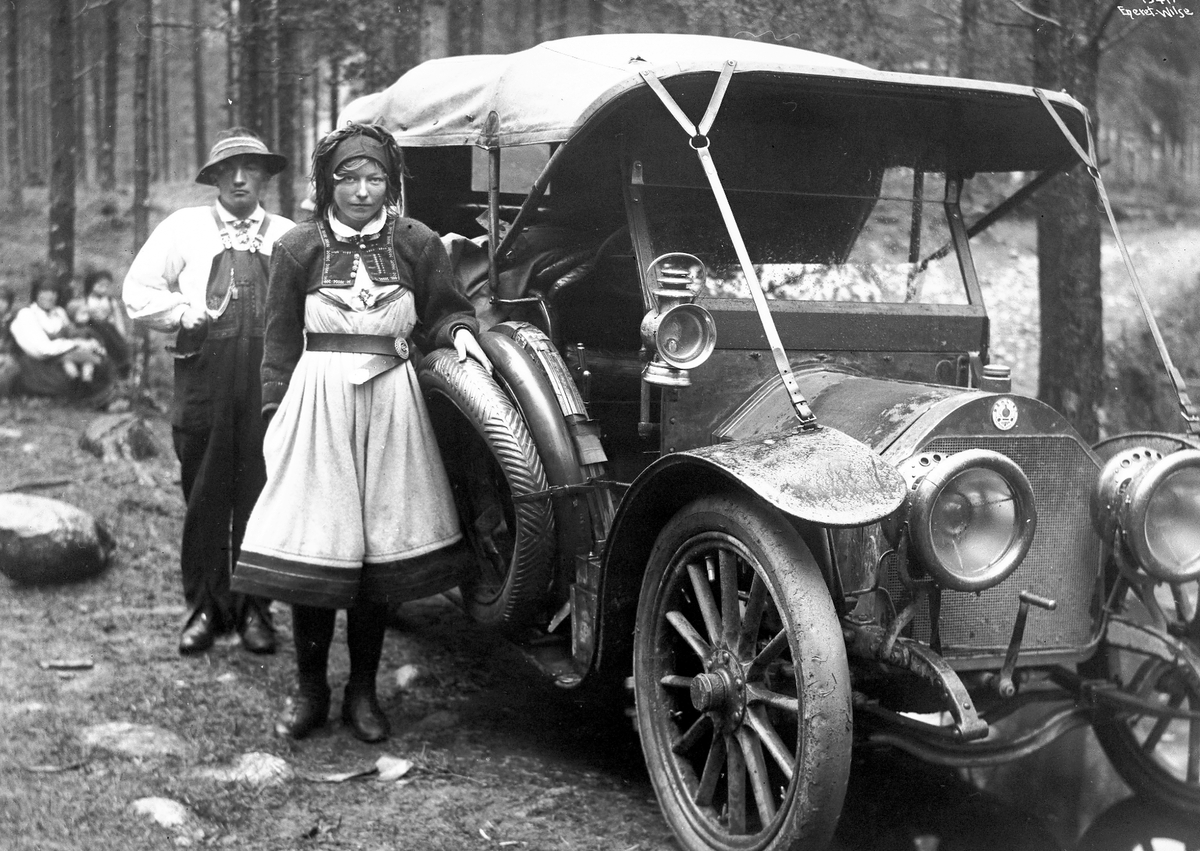 Kvinne og mann utenfor gammel bil i skogsholt.