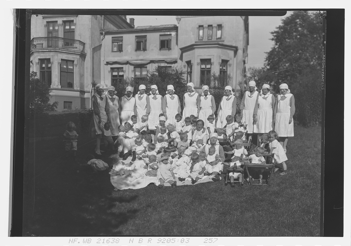 Barn og sykepleiere sitter på plenen foran Spedbarnhjemmet, Åkebergveien 23, Oslo. Fotografert 1928.