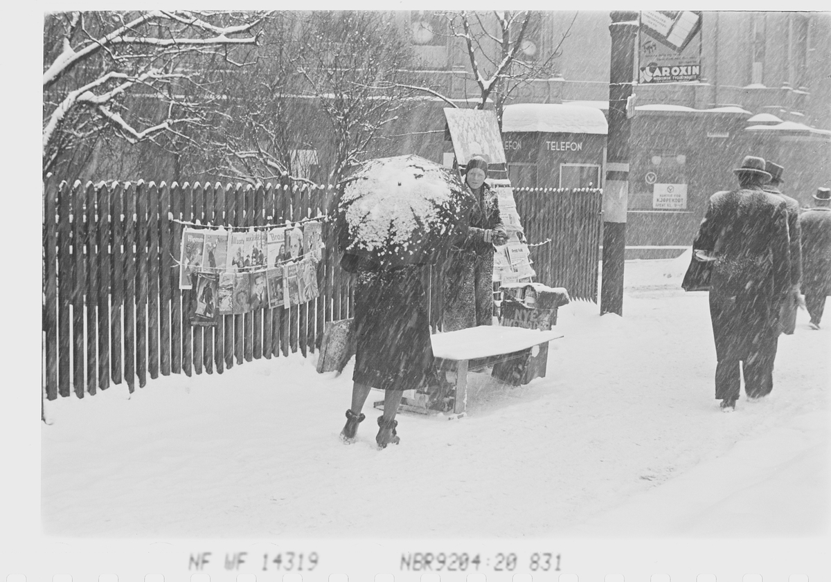 Gateselger med blader og aviser og kvinne med paraply i snøvær. Henrik Ibsens gate, tidligere Drammensveien, Oslo.
Fotografert 1941.