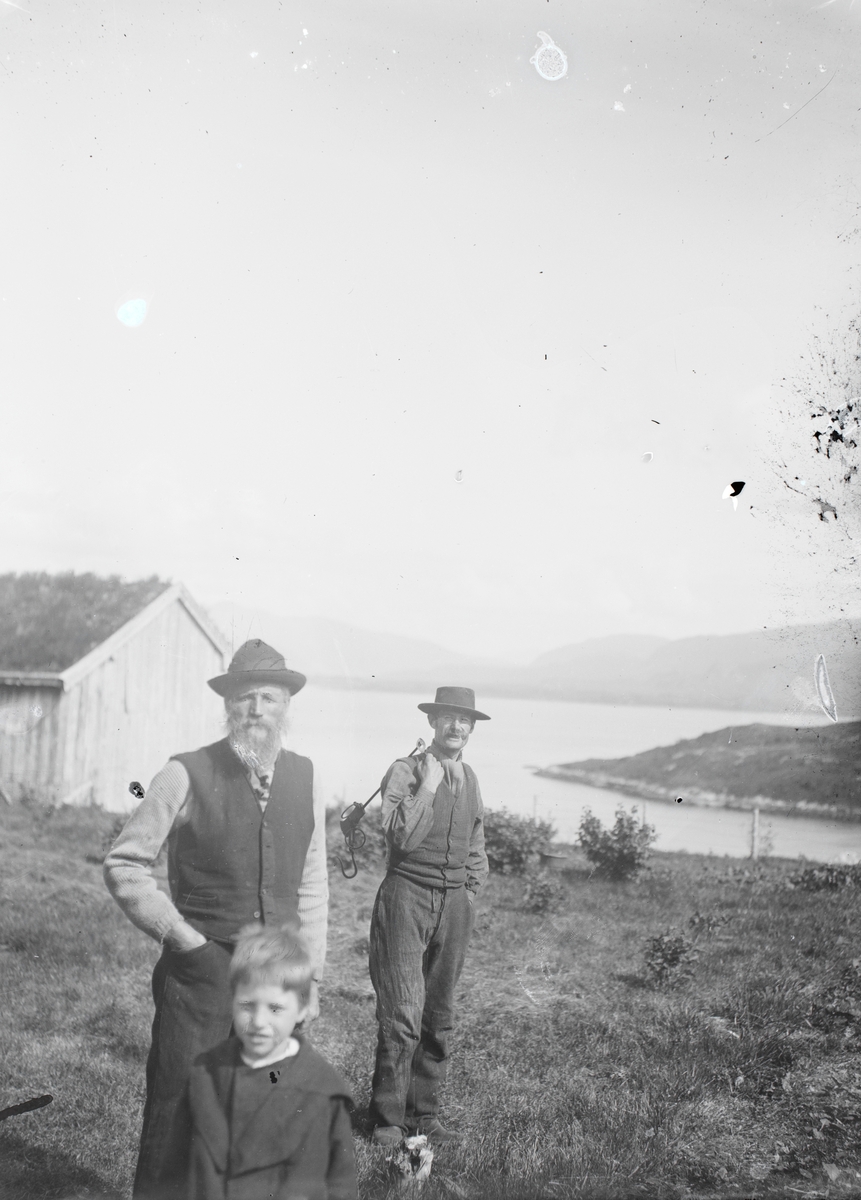 Motiv av to menn og en gutt i hagen. En av de bærer på et verktøy. Eikremsbukta i bakgrunnen.