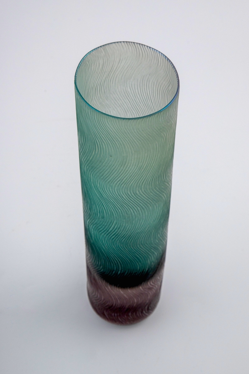 Høyreist sylinderformet vase i polykromt halvgjennomskinnelig glass. Nedre del er utført i massivt rosa glass, mens resten av vasen er grønnfarget. Yttersiden er dekorert med mattslipt dekor, i form av tette vertikale bølgende linjeføringer.