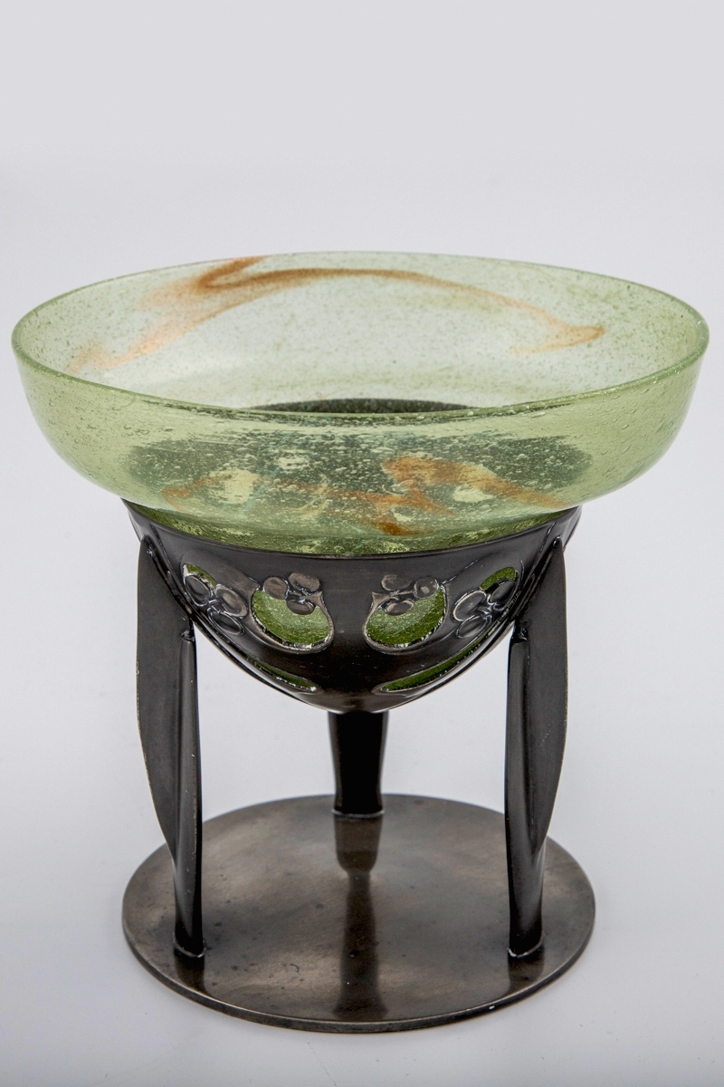 Glasskål som hviler på trebent tinnfot.  Skålen er klokkeformet med bred kant, og den eplegrønne glassmassen er fylt med små blærer. Langs kanten er det partier med innlagt bladgull. Den nedre delen bæres av en trebent fot i tinn, hvis overflate er oksidert. Den øvre delen er gjennombrutt, og smykket med stiliserte planteornamenter i lavt relieff. De tre benene hviler på sirkulær fotplate, som er noe hevet mot midten.