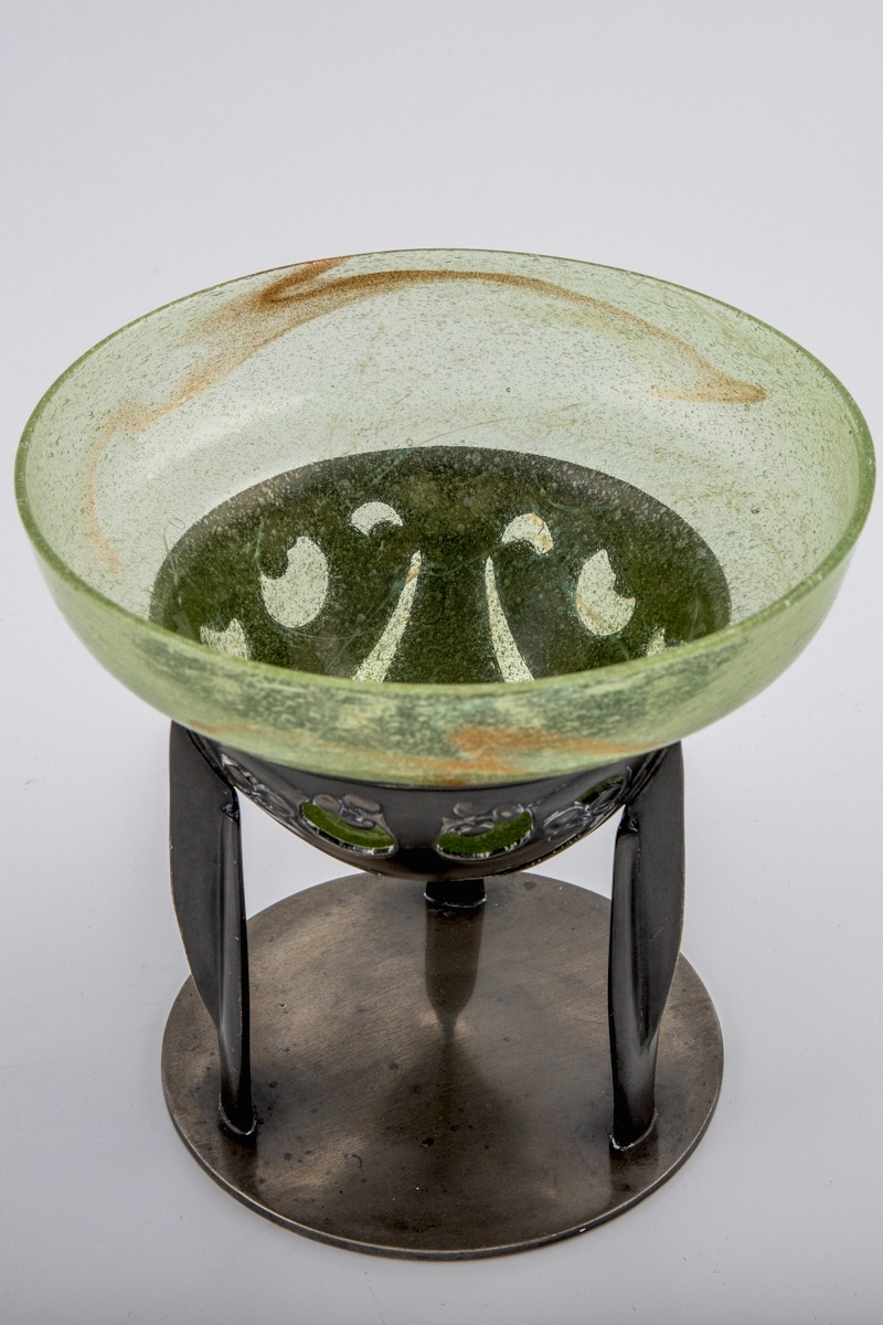 Glasskål som hviler på trebent tinnfot.  Skålen er klokkeformet med bred kant, og den eplegrønne glassmassen er fylt med små blærer. Langs kanten er det partier med innlagt bladgull. Den nedre delen bæres av en trebent fot i tinn, hvis overflate er oksidert. Den øvre delen er gjennombrutt, og smykket med stiliserte planteornamenter i lavt relieff. De tre benene hviler på sirkulær fotplate, som er noe hevet mot midten.