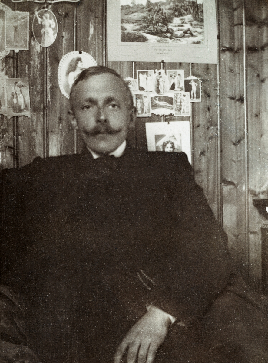Alf Herre Heltberg (f. 1878), fotografert framfor en umalt, faspanlt vegg, der det var festet en del bilder ved hjelp av tegnestifter eller nåler. Heltberg var på dette tidspunktet en middelaldrende mann med mustasje og sidekjemt hår. Han var kledd i mørke klær. Fotografiet er antakelig tatt under en jakttur. Alf Herre Heltberg var født i Løten på Hedmarken, der foreldrene, stiftskapellan Kristian Frithjof Gottlieb Heltberg og mora Kirsten Marie f. Heyerdahl var prestefolk. Seinere flyttet familien til Såset i Åsnes, der Alf gikk middelskole. Deretter flyttet han til Kristiania for å ta artium og etter hvert fortsette med videre studier. Han valgte jussen, men var også underoffiser. I 1906 giftet Alf Herre Heltberg seg med Astrid Lund fra Elverum. Svigerfaren hjalp ham å finansiere bedriften «Tenax», som skulle produsere syntetisk saft. Produktene slo ikke an, og store summer fra svigerfamilien gikk tapt. Ekteskapet ble oppløst. I 1916 giftet han seg på nytt, denne gangen med Magda Ruud fra Grefsen i Oslo. Paret flyttet til Bergen, der Alf ble leder for en meglerforretning, som også gikk konkurs. Deretter ble Alf Herre Heltberg reisende for en kjeksfabrikk. Seinere virket han som kontor- og reklamesjef og som film- og teaterhistoriker.