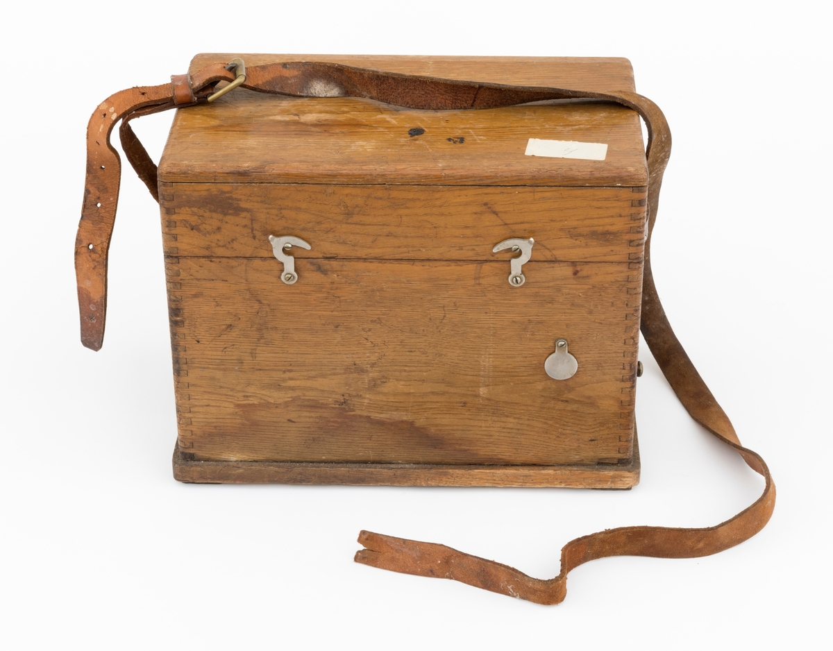 Bærbar felttelefon, antakelig av type som ble brukt tidlig på 1900-tallet. Telefonen er montert i ei trekasse, antakelig lagd av eikematerialer. Kassa har ei botnplate som er 33,1 centimeter lang, 20,2 centimeter bred og 1,5 centimeter tjukk. Ved endene av botnplatas underside er det 19,3 centimeter lange og 3,1 centimeter brede trelister som  gjenstanden sto på. Oppå den nevnte botnplata er det ei 32 centimeter lang, 19 centimeter bred og 16,6 centimeter høy kasse med fingerskjøtte, noe avrundete hjørner. Til denne kassas bakre langside er det hengslet et lokk av samme type materiale og med samme lengde og bredde som kassa. Lokket er 6,1 centimeter høyt. Lokket stenges ved hjelp av dreibare kroker på kassas frontside som vendes, slik at de omslutter skruer på lokkets frontside. Litt lengre neden på kassefronten er det et gjennombrutt hull med 1,3 centimeters diameter, antakelig innføringspunktet for sveiva til telefonen. Dette hukket kan stenges ved hjelp av ei lita, dreibar stålskive når sveiva ikke er i bruk. Da festes sveiva i en trekloss som er innlimt i et hjørne i lokket. Telefonen bæres i ei cirka 2,4 centimeter bred lysebrun lærrein, som er festet i messingknapper, sentralt på den nedre delen av kassas tverrender. At kassa bæres med den riktige sida opp er sikret ved hjelp av smøyestoler av lær høyere oppe på de samme endeflatene. Lengden på bærereima kan justeres ved hjelp av ei nålespenne. Inne i kassa er det antakelig et batteri, skjult under trelokk i to plan. På det nedre er det inntappet en toleddet, forkrommet holder for telefonrøret. Også telefonrøret er forkrommet, med har bakelittkomponenter for håndgrep (midten), øreanlegg og taletrakt. Sentralt på lokkets underside er det pålimt en trekloss med et tjukt svart filtbelegg, som skulle holde telefonrøret på plass under transport. Ellers er det innlimt to etiketter på undersida av lokket. Den ene er et firmamerke som refererer til «AKTIESELSKABET ELEKTISK BUREAU, KRISTIANIA, NORGE». På dette merket er det et rødt skjold med en løvefigur med krone over. Undersida av lokket er det også påklistrert en etikett med tekniske spesifikasjoner fra produsenten. Denne kontrollseddelen har også vært nummert, datert og signert, men hjørnet hvor blant annet årstallet har stått, er dessverre avrevet.