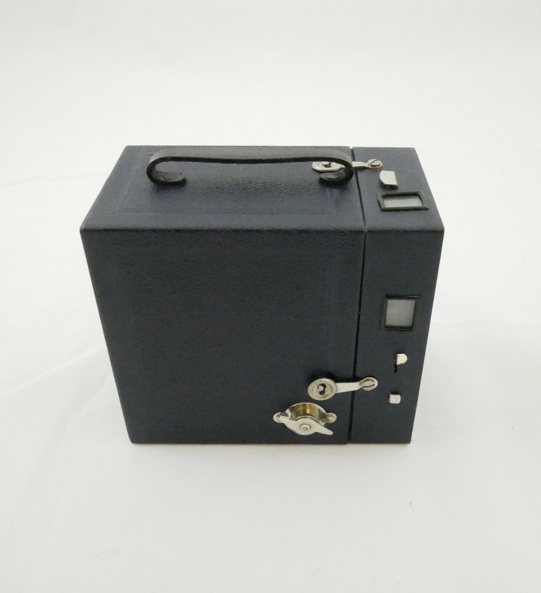 Brun pappemballasje som kan åpnes i den ene siden. Emballasjen er festet med stifter i den ene enden. På den ene kortsiden er det påklistret en merkelapp med blå bakgrunn og bokstaver i gullfarge.

Påskrift: 
Metall // Roll- Film // BOX- CAMERA // 6 ½ X 11 - 2 ½ X 4 ​​​1⁄4 // MENISKUS // Foc. 1 : 11 // Nr. 160

Inne i emballasjen er det et kamera. Kameraet er inne i et etui av blått lær, på toppen er det et svart håndtak i samme materiale. Etuiet kan åpnes i den ene siden og er festet med to metallhekter. 