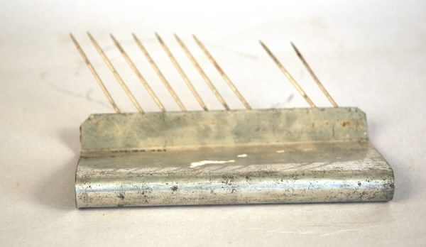 Kakstöd av metall, som används som stöd vid perforering av sega kakor med cellperforator. Hopvikt metallskiva som avslutas i ett veck i vilket vassa metallnålar är fastsatta. Saknas: tre nålar.