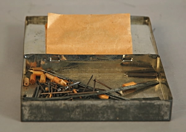 Rektangulär ask av plåt med lock på gångjärn. Tre små hål i locket. Inuti asken finns ett antal avståndshylsor av trä, spik, 5 tunna metallremsor samt två vinklade specialskruvar i en papperspåse.
