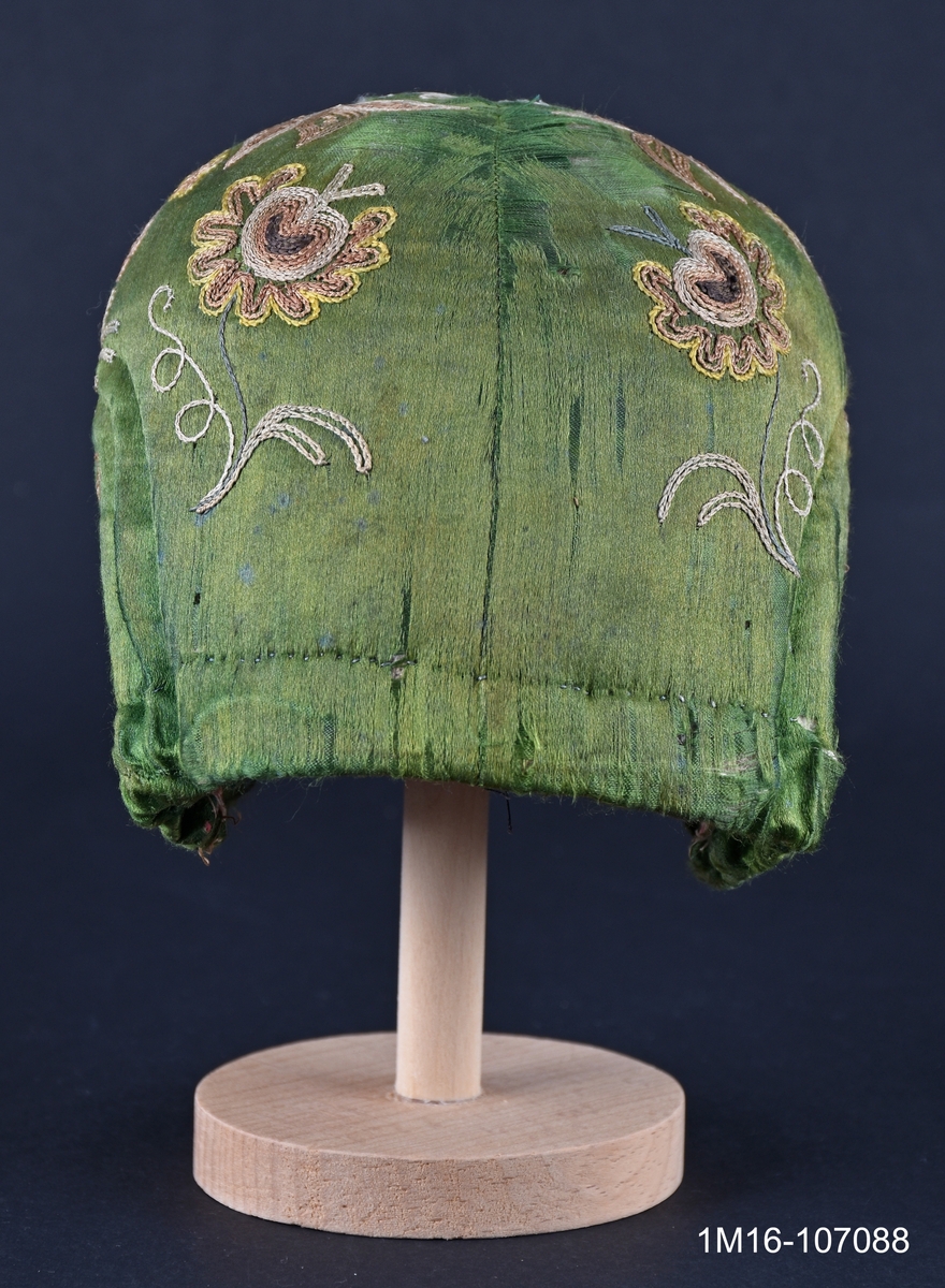 Bindmössa av grönt siden med gulbrunt tambursömsbroderi i bruna nyanser. Bindmössan kopierades till Valledräkten under 1960-talet av ett dräktråd. Har tillhört Carolina Johansson (1835-1917).