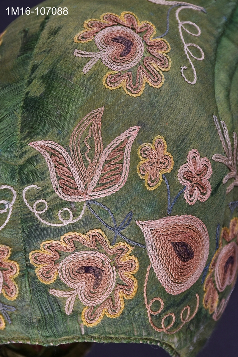 Bindmössa av grönt siden med gulbrunt tambursömsbroderi i bruna nyanser. Bindmössan kopierades till Valledräkten under 1960-talet av ett dräktråd. Har tillhört Carolina Johansson (1835-1917).