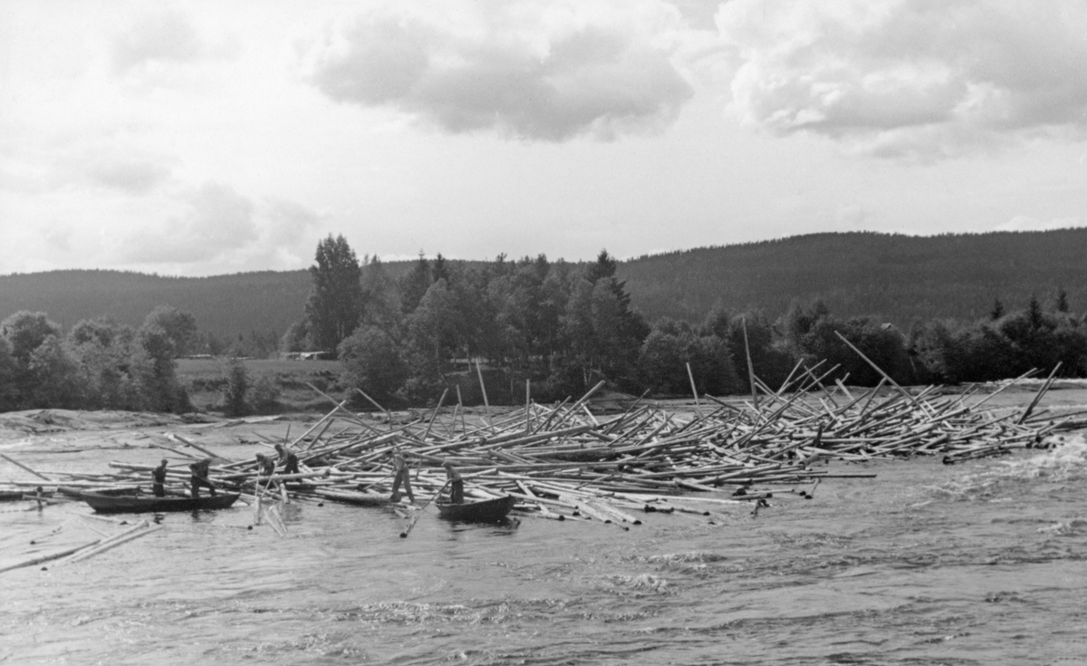 Fra Gjølstadfossen i Brandval, cirka ni kilometer nord for Kongsvinger by.  Fotografiet er tatt fra den østre elvebredden mot en tømmervase som har samlet seg opp i fossestrykene om lag midt i elveleiet.  Vi ser hvordan fløterne har rodd opp mot den nedre delen av tømmervasen og startet arbeidet med arbeide ut stokkene.  To karer befinner seg i en båt, en i en annen, mens til sammen tre arbeider fra kanten av tømmervasen og med kort avstand til de to båtene.  Langs den vestre elvebredden bak fløterne og tømmervasen vokste det et belte av lauvskog.  På slettelandet bakenfor skimter vi flatt åkerland og helt i bakgrunnen møter en skogdekt åsrygg den lettskyete himmelen.  Dette bildet ble tatt i midten av juli 1941.  