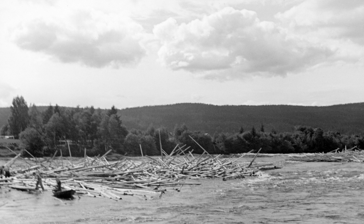 Fra Gjølstadfossen i Brandval, cirka ni kilometer nord for Kongsvinger by. Fotografiet er tatt fra den østre elvebredden, skrått i motstrøms retning mot to tømmervaser som har samlet seg opp i fossestrykene om lag midt i elveleiet.  Lengst til venstre i dette bildet ser vi hvordan fløterne har rodd opp mot den nedre delen av den nederste tømmervasen og startet arbeidet med arbeide ut stokkene.  Vi ser en mann i en robåt like inntil haugen av tømmerstokker.  Like ved båten arbeider en mann med fløterhaker.  Noe lengre unna (til venstre) skimter vi også to andre fløtere med haker, som antakelig hadde sikring i form av en robåt som lå utenfor venstre bildekant. Langs den vestre elvebredden, bak fløterne og tømmervasen, vokste det et belte av lauvskog.  Bakenfor aner vi et sletteland før en skogdekt åsrygg hever landskapet opp mot den lettskyete himmelen.  Dette bildet ble tatt i midten av juli 1941.