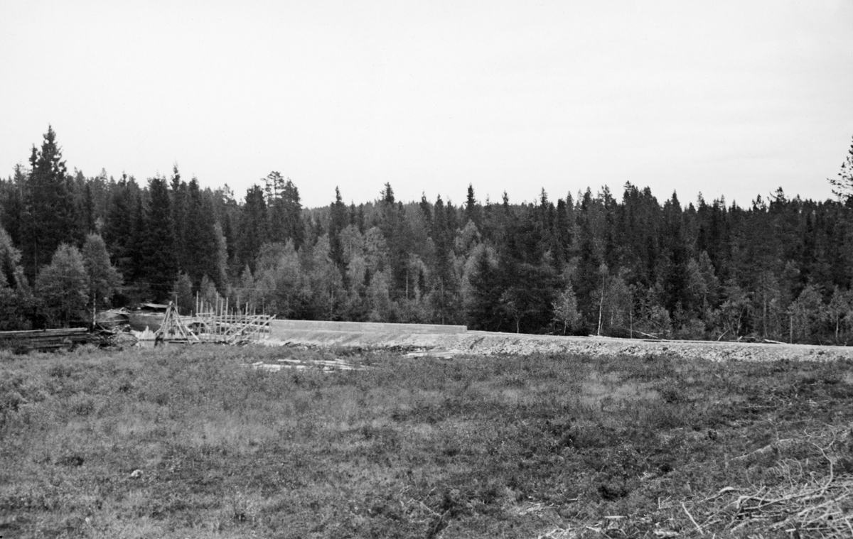 Fra bygginga av den nye Storsjøseterdammen i Kynnavassdraget høsten 1941. Fotografiet er tatt fra utkanten av det som skulle bli damområdet, men som på dette tidspunktet var ei grasbevokst flate med svak skråning ned mot vassdraget.  I forgrunnen lå det en del kvist.  I mellomgrunnen er det opplagt en lang damarm av grus.  Der denne grusryggen møter vassdraget skimtes temporære trekonstruksjoner, antakelig stillaser for bygging av damåpningen med omliggende steinkar.  Bak damarmen og på motsatt side av vassdraget vokste det granskog med spredte innslag av lauvtrær. 