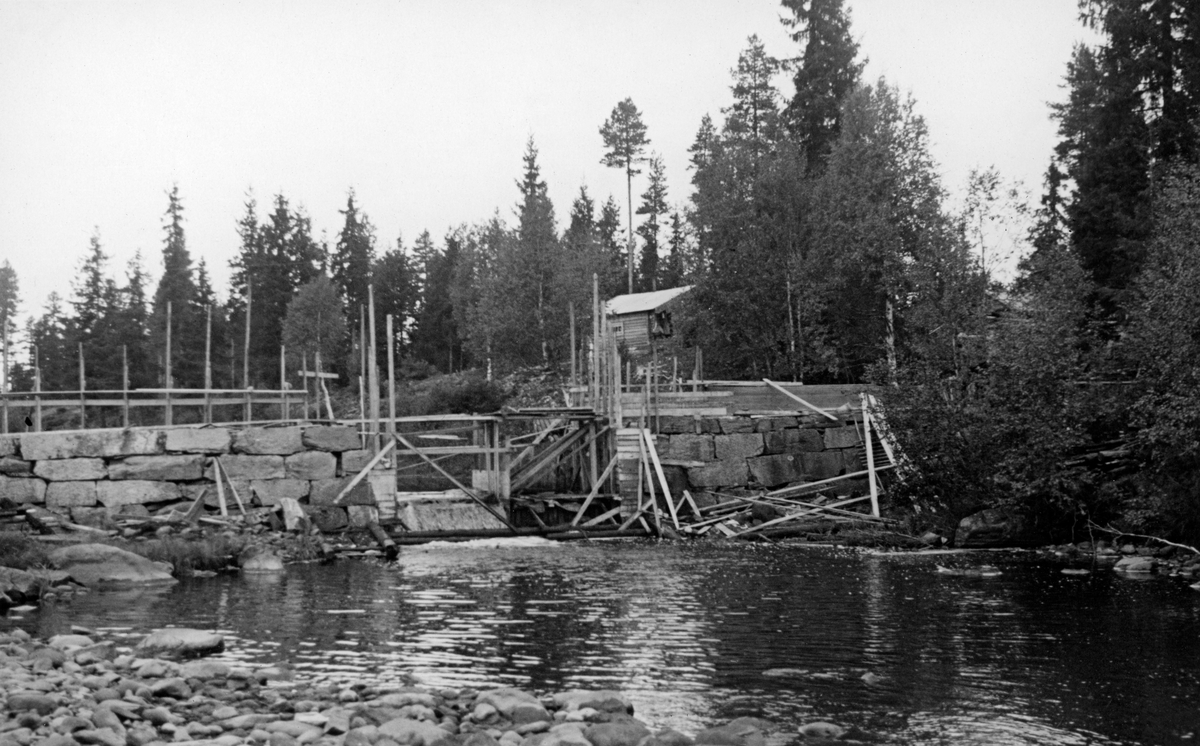 Den nye Storsjøseterdammen i Kynnavassdraget, fotografert under byggeprosessen høsten 1941.  Bildet er tatt fra motstrøms side med kulpen nedunder dammen i forgrunnen.  Nedsida av damarmene, på begge sider av åpningen, er murt i grov, men tilhogd gråstein.  På baksida av damarmene og i løpet ser vi deler av temporære trekonstruksjoner, antakelig stillaser.  En tett bordskjerm på damkrona til høyre kan minne om ei forskaling til en støpt konstruksjon.  På en bakkekam bakenfor dette igjen skimtes damkoia, ei laftet hytte med inngang på gavlveggen.  Grana later til å ha vært dominerende treslag i området, sjøl om det også finnes furu, bjørk og muligens or (mot elveløpet). 