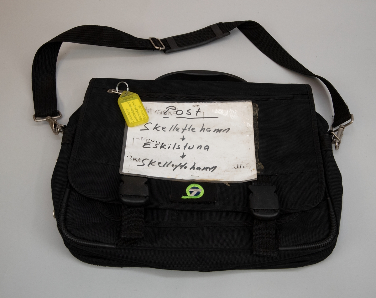 Postväska för internpost i form av en svart portfölj med en fastlimmad broderad logotyp för TGOJ trafik AB och en fastlimmad och fasthäftad plastficka med ett vitt pappersark med texten "Post Skelleftehamn -> Eskilstuna -> Skelleftehamn". "Post" är understruket. Det finns också spår av en tidigare tryckt text på plastfickan som bör ha varit densamma "Postväska...skilstu...Ske...una". Väskan har en axelrem och bärhandtag. I dragtappen till blixtlåset på en ficka i locket sitter en nyckelring med en gul nyckelbricka av plast med en etikett med tryckt text "Eskilstuna - Skelleftehamn / Skelleftehamn - Eskilstuna".