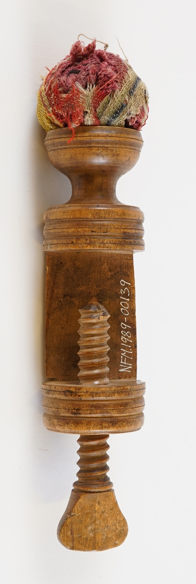 Ei syskrue i tre, med pute i tekstil på toppen. Nytta til å setje fast arbeidet til bordet når ein sydde.