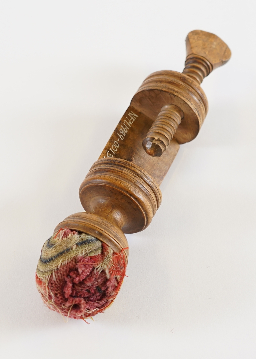 Ei syskrue i tre, med pute i tekstil på toppen. Nytta til å setje fast arbeidet til bordet når ein sydde.