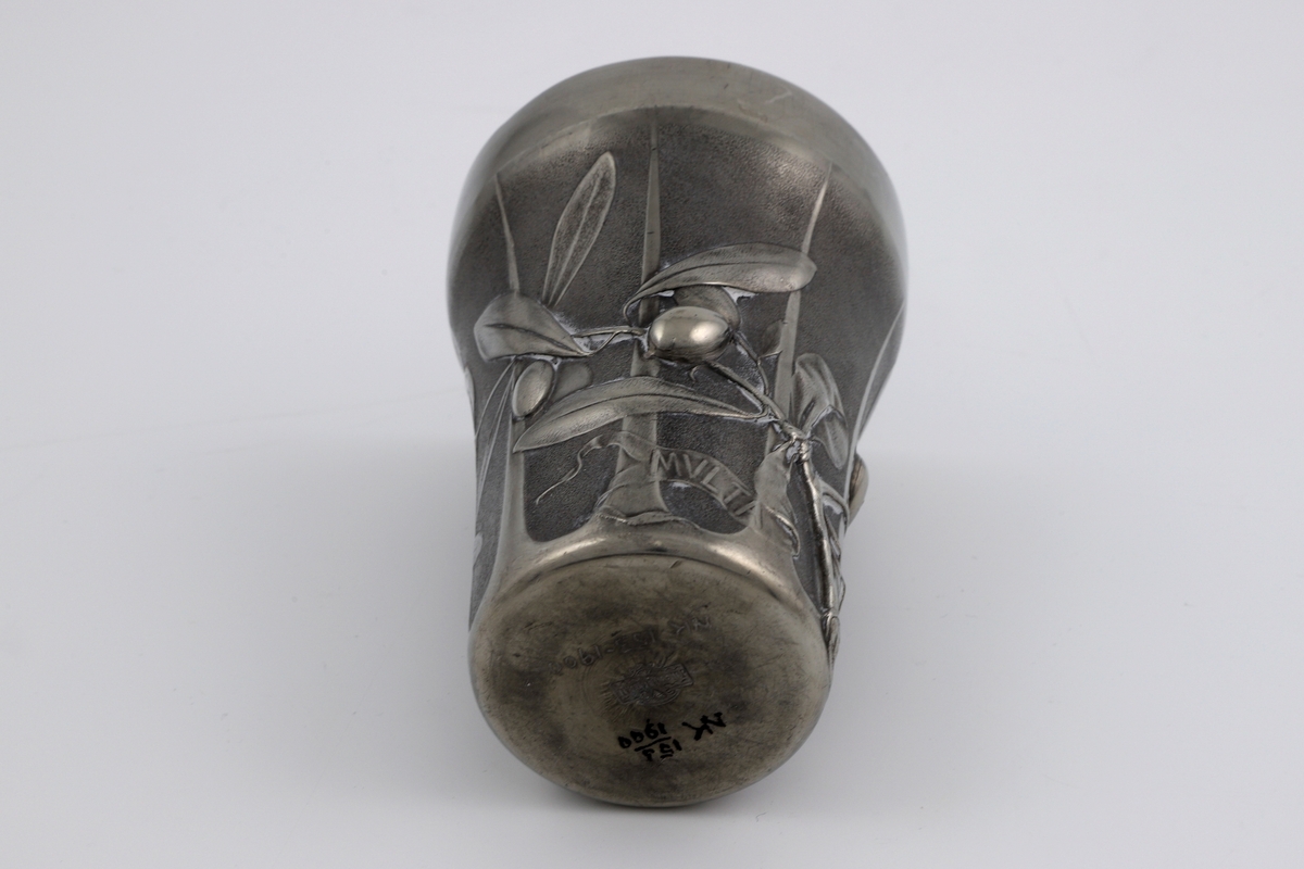 Beger i tinn. Formen smalner svakt nedover. Dekoren på korpus viser en olivenkvist i lavt relieff, derav begerets tittel L’Olivier (fr. oliventre). Til grenen er det festet to bånd med innskriften «MVLTA PAVCIS» (lat. mye med lite). Kanten er blank og glatt, mens korpus trolig er patinert. Yttersiden er inndelt med gjentagende rifler.