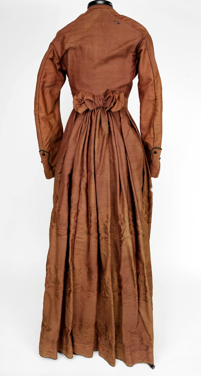 Todelt kjole i tynn brokade med blomstermønster innvevd på skjørtet. Kjolelivet er helfòret med to ulike stoffer. ermene er sydd i for hånd og sårkantene kastet over for hånd. stikningssømmer er sydd på maskin. Sidesømmer og innsnitt med lett spiling likeså. Kjolelivet lukkes foran med stofftrukne knapper og to hekter nederst. Nederst på ermene er det pyntet med oppbrett med grønne bånd, kapper og en effekt i kjolens stoff. Ermene er skåret i to deler.
Skjørtet er foldet foran med splitt i fold på venstre side som lukkes med hekter på to steder. Nederst på innsiden er det skoningskant og slitesnor. Bak i livet er det et felt på 6 cm med striping.
Belte med hektelukning under sløyfe som har sittet bak.
Kjøpt stoff. Muligens brukt som modell for stakk til Sør-Østerdalsbunaden.