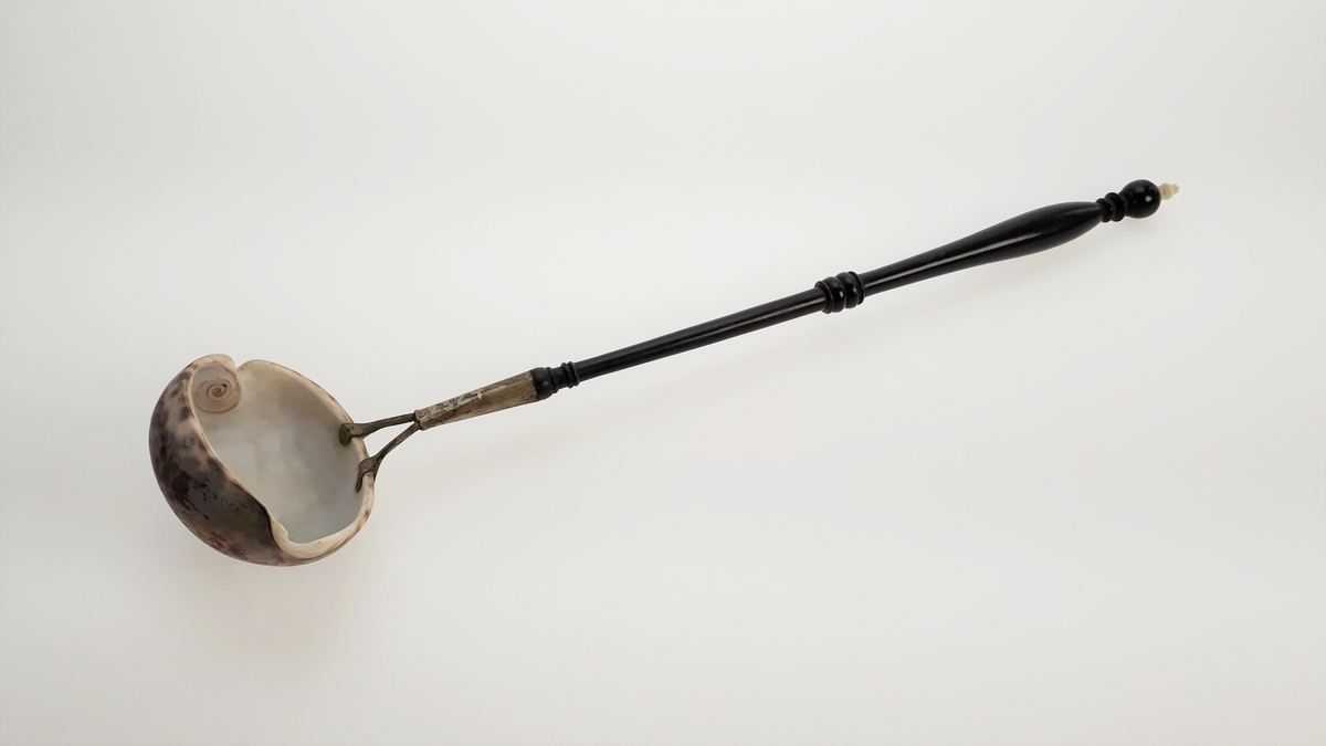 Suppeøse med dreid håndtak i tre og øseskål av skjell. Skjellet er festet til skaftet med en metallhylse som er naglet fast i skjellet. På toppen av skaftet er det en liten dreid knott. Denne er antagelig av elfenben.