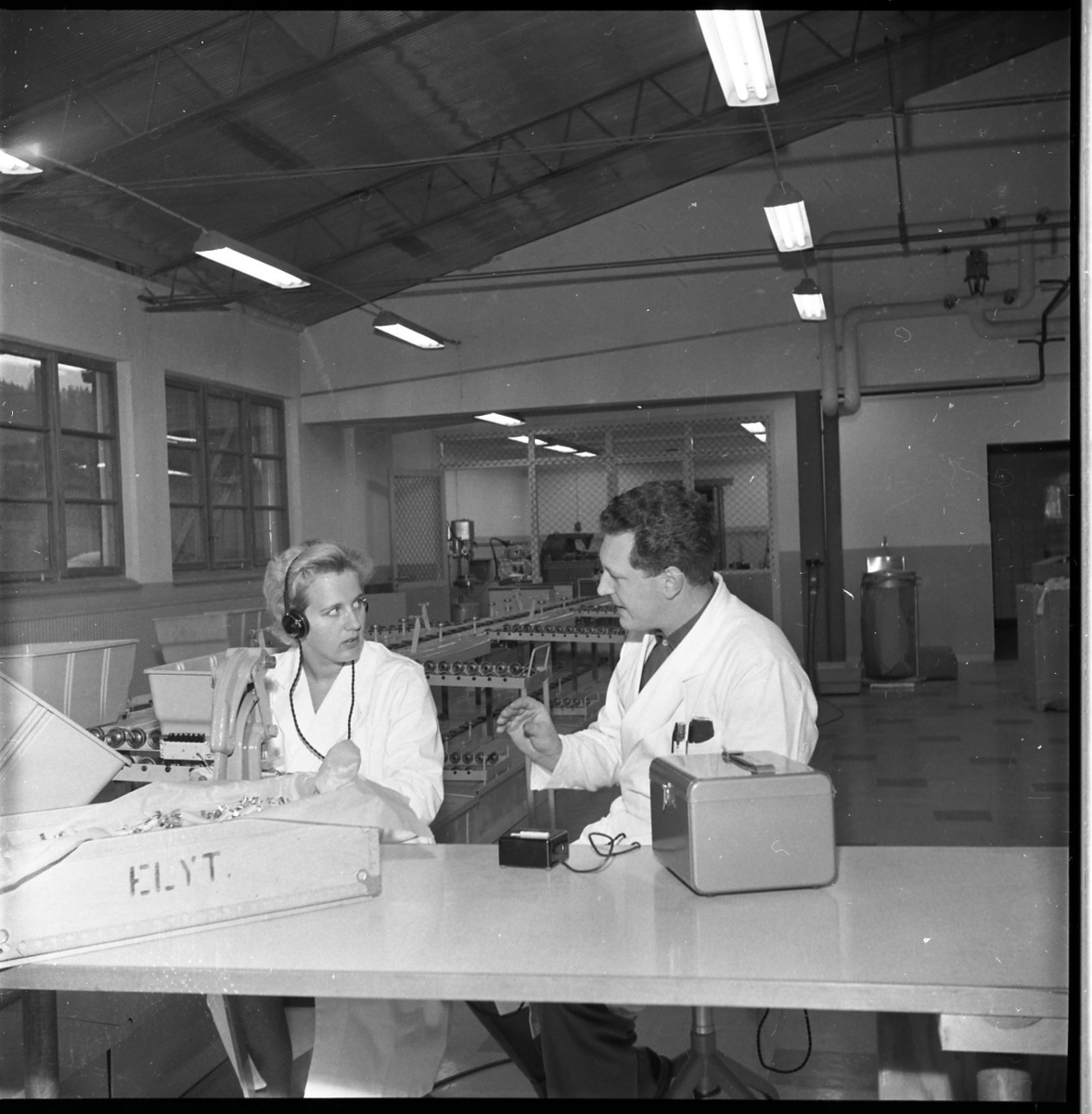 En kvinna och en man i vit arbetsrock sitter vid en arbetsstation på Rifa-fabriken i Gränna. Kvinnan har öronskydd/hörlurar. De är Inga Johansson och Göran Andersson.