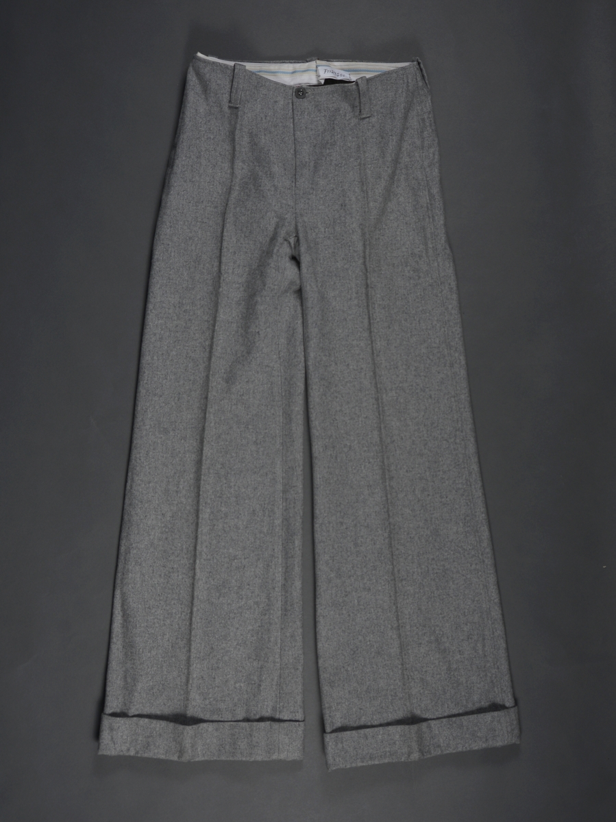 Slengbukse i grå flanell. Enkel modell uten linning men med beltestropper. Gylfåpning og knapp foran. Sleng i buksebeina, oppbrett nederst.