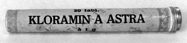 "Tablettrör av lättmetall. Rör med flat botten och skruvlock. Gul botten med ihuvudsak svart text: ""20 tabl. KLORAMIN A ASTRA Ã  1 g"". Bruksanvisningfinns också på röret. Handskrivet: ""1 06"". Vid ändarna är röret dekorerat i röttoch vitt."
