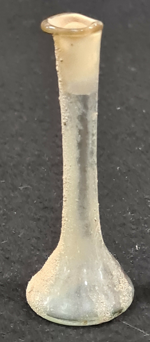 Glaskärl med 7,5 cm. hög, ca 1 cm . tjock, hals vilken är något ''utfluten'' vid mynningen. Själva kärlkroppen har en rund botten och upp mot halsen en avsmalnande form. Glaset är klart men på en sida täckt av en grå, sandliknande, beläggning.

Enl. uppgift av givaren är kärlet  ett så kallat ''tågglas'' från den romerkas staden pompeji. Den sandliknande beläggningen skall vara vulkanaska från Vesuvius. Ekholm fick kärlet i gåva av en antikhandlare från Belgrad. Enl. denne antikhandlare användes tårglaset vid begravning för att samla upp de sörjandes tårar, i varefter glaset nedlades med den döde i graven.