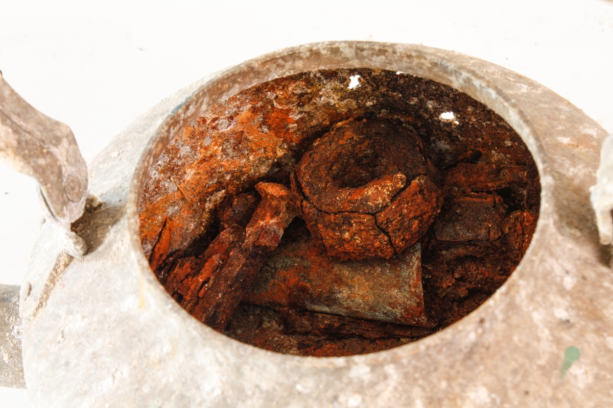 Kaffekjele i hvitt metall med helletut og hengslet håndtak. Hvert hengsel er festet til kroppen med to nagler, og til håndtaket med én nagle. Håndtaket er ikke lenger bevegelig, og står rett opp. Helletuten er festet nederst på kroppen, og er svakt avsmalnet og bøyd opp og utover. Kjelen mangler lokk. Kjeleformen er konisk med innsvinget topp. Metallet er matt, med flere ujevne korrosjonshull i den ene siden. Det er små sorte, hvite og grønne malingsflekker på overflaten.

I kjelen ligger diverse små gjenstander.
- 24 jernspiker i ulike størrelser, flere er trolig belagt med sink.
- En ringklemme med skruegjenger, trolig jern belagt med sink.
- En trekork.
- Tre små jernbiter, uidentifisert.
- Tre ringer/deler av ringer, av jern og kobber.
- Fire jernmuttere, ulike størrelser.
- En blålig taustump, trolig bomull.
- En jernsylinder med skruegjenger.
- En skruebolt av jern.
- En kobbersylinder med hull i sidene og et buet jernlokk i en ende.
- Flere fastrustede spiker og muttere.
