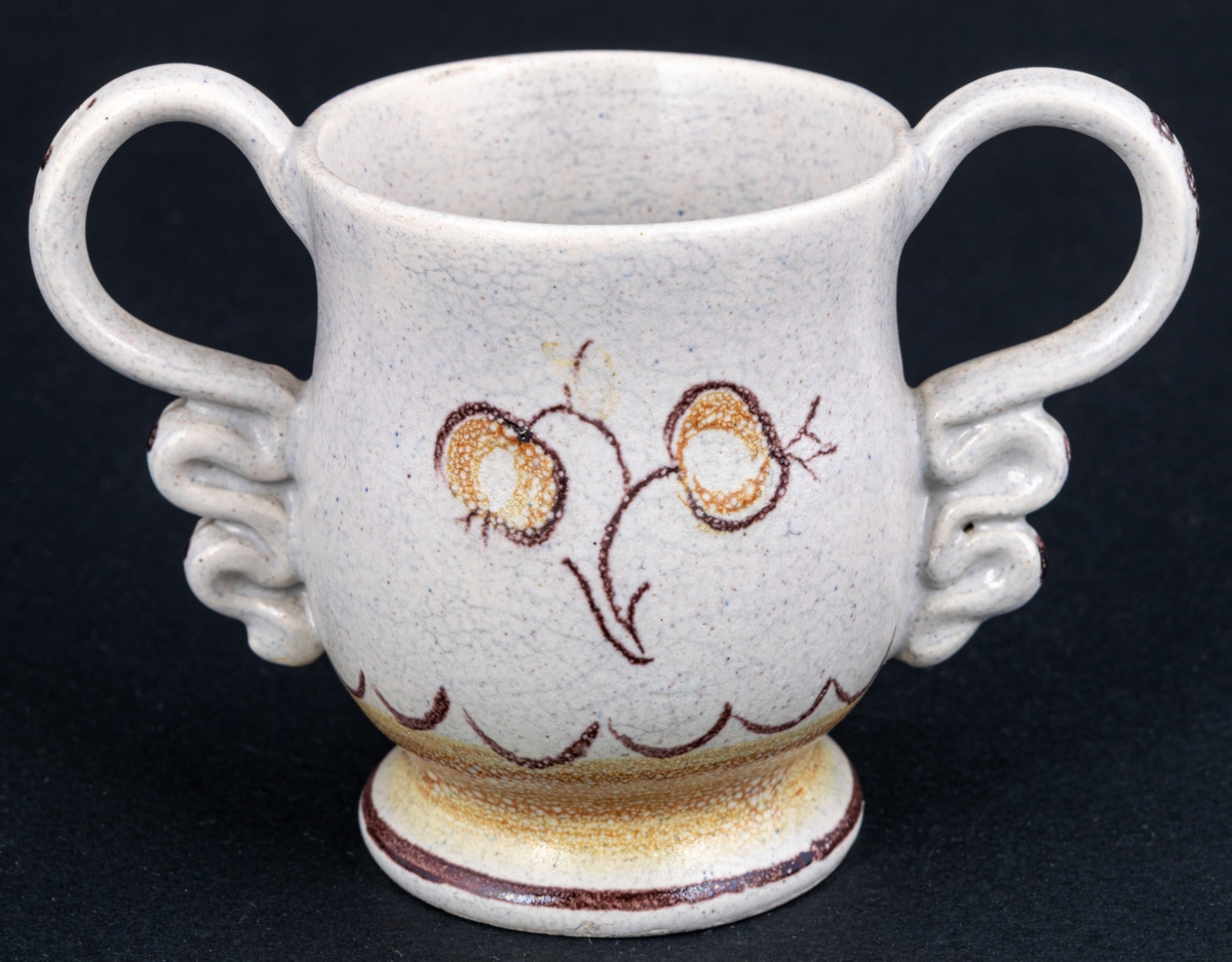 Bägare av lergods. Bo Fajans, design Eva Jancke­Björk. Grå S­svängd med stora hänklar, ljus- och mörkbruna partier nedtill samt dekor av en blomma.