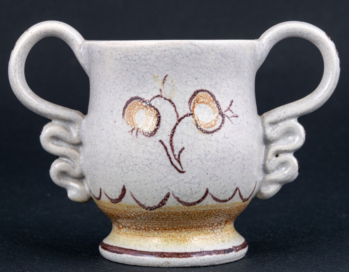 Bägare av lergods. Bo Fajans, design Eva Jancke­Björk. Grå S­svängd med stora hänklar, ljus- och mörkbruna partier nedtill samt dekor av en blomma.